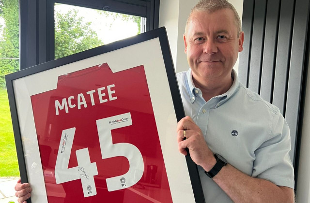 Man stood in front of window, smiling, wearing a blue shirt.  He is holding a framed, Barnsley football shirt.