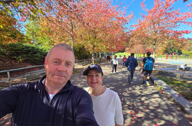 Man and woman stood in a park, with autumnal trees behind them, of a red and orange colour