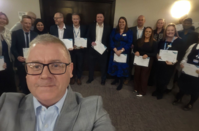 Man stood in front of a large group of people, holding certificates