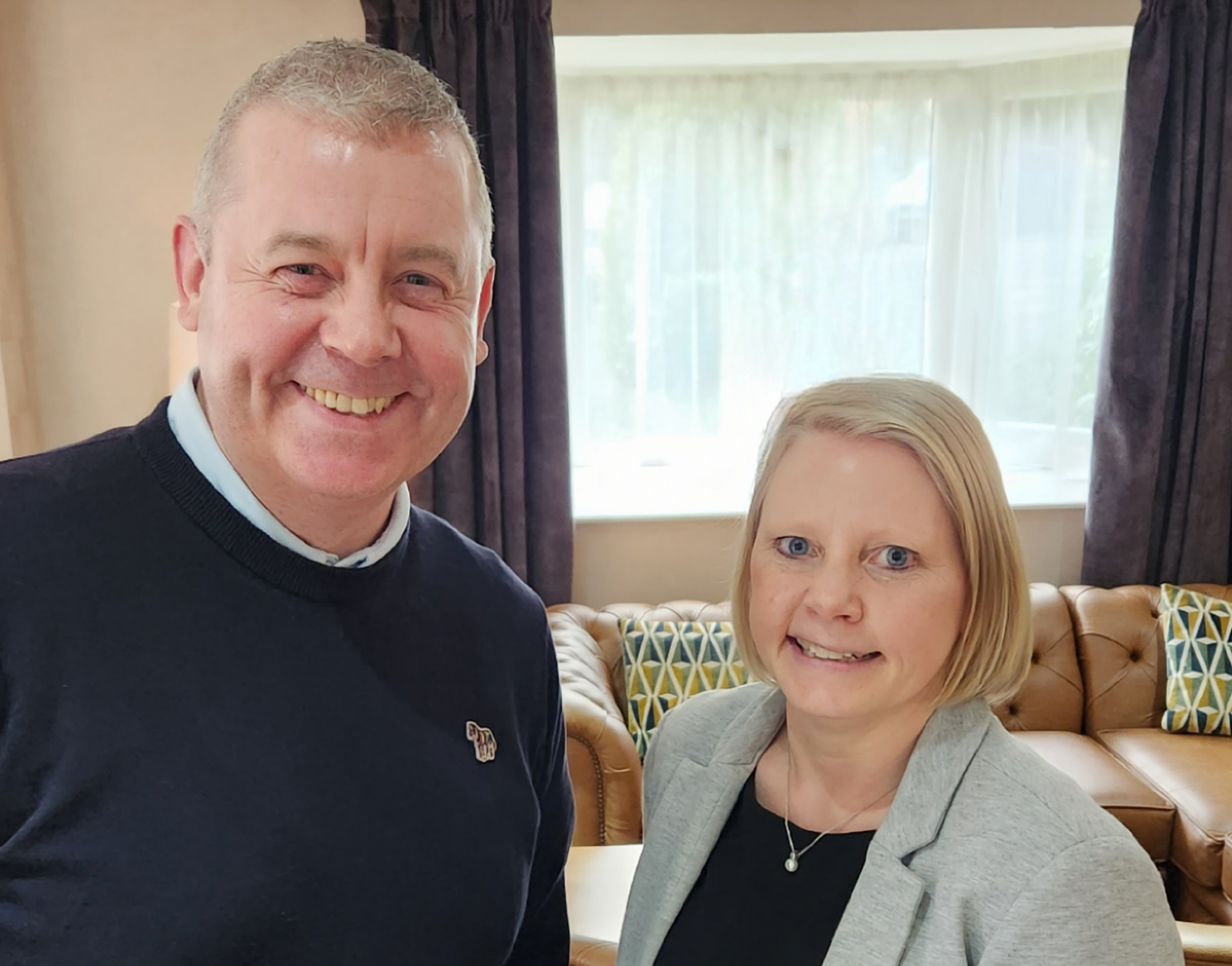 A man in a blue jumper smiling, with a women with blond hair, blue eyes, wearing a gray jacket smiling