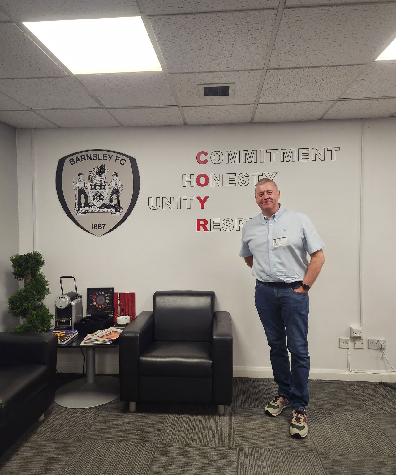 Man stood next to a chair, in front of a sign reading 'Commitment, Honesty, Unity, Respect'.