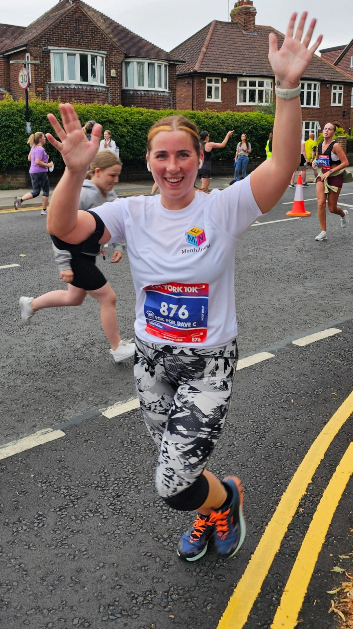 A woman running the York 10k, with her arms in the air in celebration.  She's wearing a white t-shirt, with the manfulness logo on.