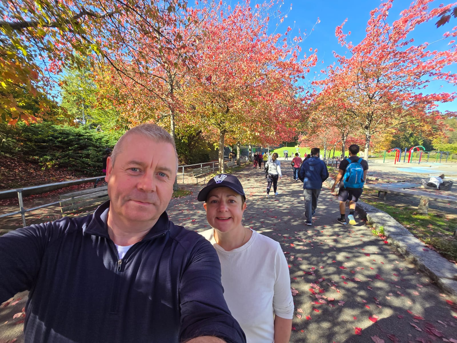 Man and woman stood in a park, with autumnal trees behind them, of a red and orange colour