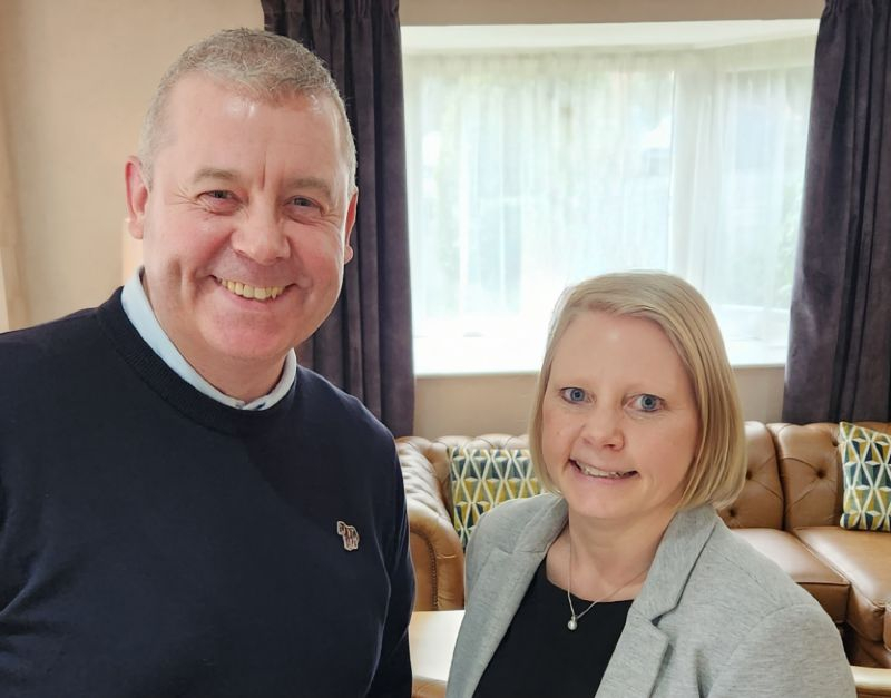 A man in a blue jumper, stood next to a woman in a grey jacket.  Both are stood in front of a sofa and a window.